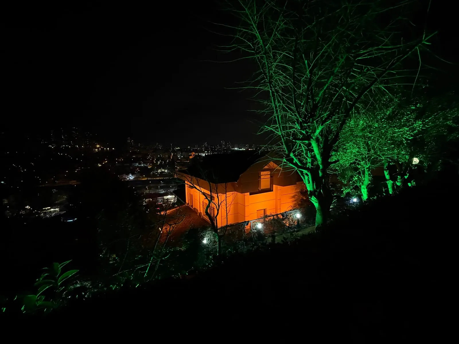 Yamaçtaki evin turuncu cephe aydınlatması ve yeşil ağaç ışıkları, gece