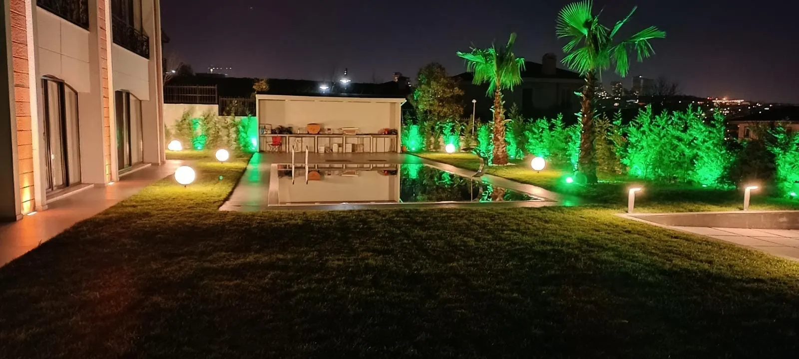 Havuzlu villa bahçesinde gece: palmiyeler, yeşil ağaç aydınlatması ve yol armatürleri