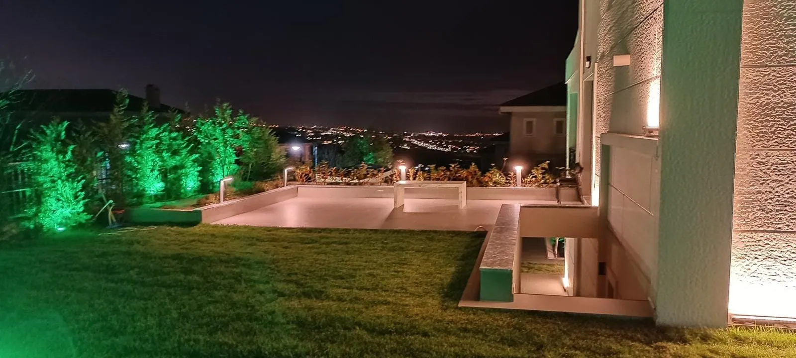 Havuz kenarı ve bahçe sınırındaki bitkilerin yeşil aydınlatması, arkada şehir ışıkları